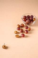 Scattered sliced half ripe sweet plum fruits with water drops near to plums in glass bowl on cream colored background, bright, angle view, selective focus