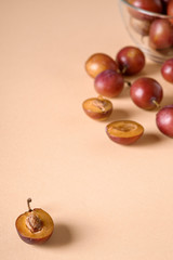 Scattered sliced half ripe sweet plum fruits with water drops near to plums in glass bowl on cream colored background, bright, angle view, selective focus