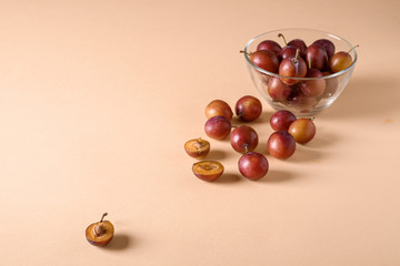 Scattered sliced half ripe sweet plum fruits with water drops near to plums in glass bowl on cream colored background, bright, angle view, selective focus