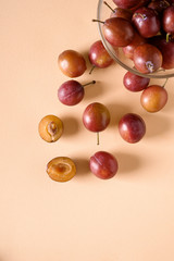 Scattered sliced half ripe sweet plum fruits with water drops near to plums in glass bowl on cream colored background, bright, top view, flat lay