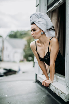 Woman just with a towel in her hair leans out a window while dressing