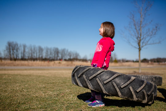 A strong little girl struggles to hold up a large tire in a park