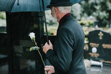 Elegant sad elderly man standing on the rain with umbrella and grieves at the grave of a loved...