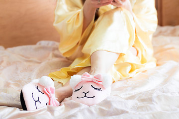 Fototapeta na wymiar female feet wearing cute llama trendy pink slippers, lay on bed with wooden headboard and on pastel silk gown. Home Rest Relax Holidays Horizontal