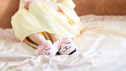 Fototapeta na wymiar female feet wearing cute llama trendy pink slippers, lay on bed with wooden headboard and on pastel silk gown. Home Rest Relax Holidays Horizontal with copy space