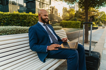 Fototapeta premium Indian businessman holding laptop notebook wearing blue suit near office. Freelance business work concept. His suitcase is near bench