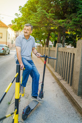 Senior man sitting on the fence by the sidewalk holding electric kick push scooter in a summer day