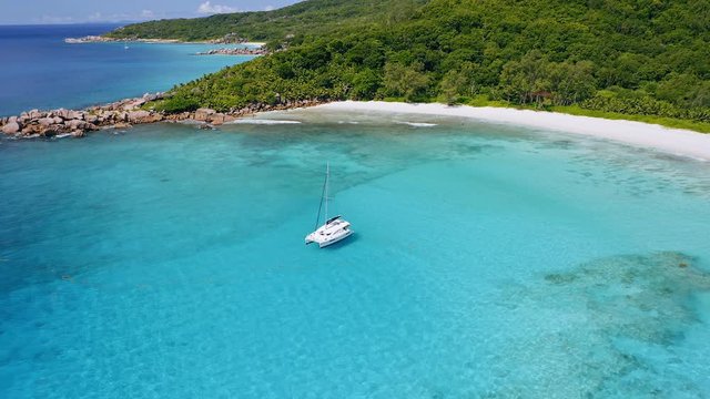 Aerial view of luxury catamaran yacht moored in crystal clear blue lagoon. Amazing tropical sandy beach surrounded by mountain ridge and rainforest in background. La Digue island, Seychelles