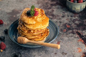 Delicious pancake with blueberries, raspberries and honey in blue dish on rust table. Still life in rustic style.