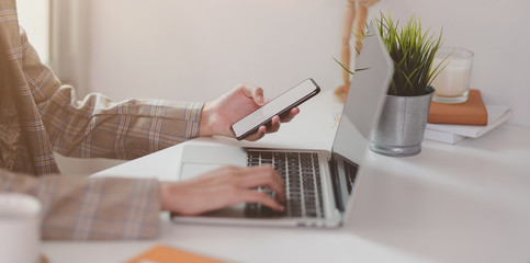 Fototapeta premium Young businesswoman typing on laptop while holding smartphone
