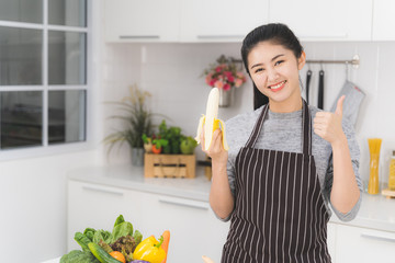 Portrait of asian beautiful woman and housewife is smiling and holding banana in white kitchen. Healthy food concept. © amornchaijj