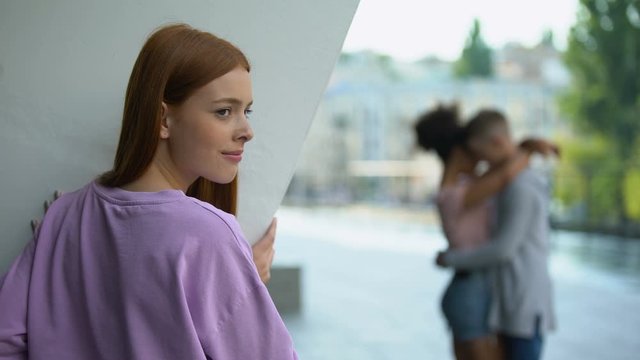 Unhappy female teenager looking at male classmate hugging girlfriend, jealousy