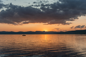 Burning red sunset on the river. Men riding boat. Silhouette of mountains on the background. Beautiful clouds in the sky.