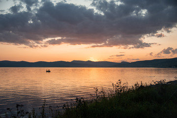 Burning red sunset on the river. Men riding boat. Silhouette of mountains on the background. Beautiful clouds in the sky.