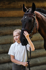 Obraz premium A teenage girl jockey stands next to a brown horse and hugs her. Against the background of a wooden wall stables.