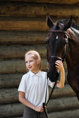 Obraz premium A teenage girl jockey stands next to a brown horse and hugs her. Against the background of a wooden wall stables.