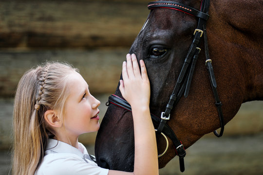 A teenage girl jockey stands next to a brown horse and hugs her. Against the background of a wooden wall stables. - Powered by Adobe