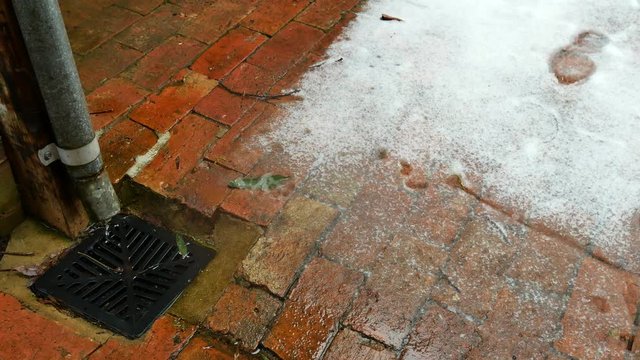 Water flows from a downpipe into a drain with a brick path and snow.