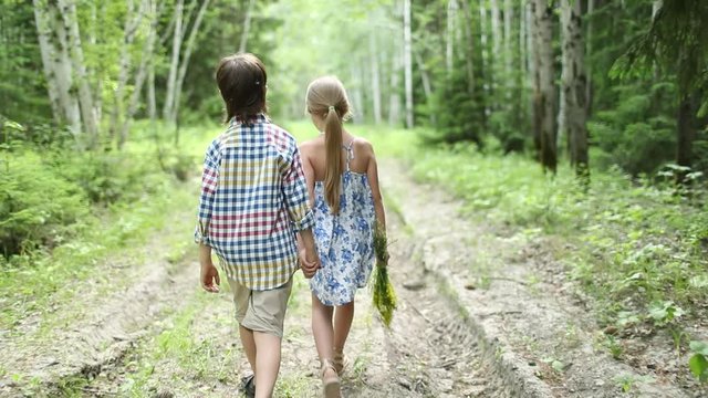 A boy and a girl with a bouquet are on the road in the woods holding hands. The view from the back. Children, nature, summer casual fashion. Beauty. First love.