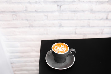One gray cup of fresh cappuccino on black table. Background of white textured brick wall, loft style interior. Black and white minimalist style. Empty place for text, copy space.