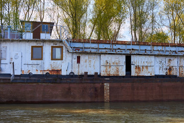 Old broken damaged rusty metal river barge on near the shore with green trees at early spring