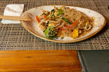 Asian dish - stewed noodles with vegetables, seeds and fruits on a white plate, the edges of which are sprinkled with spices.