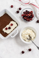 Homemade brownie cake preparation, chocolate dough with cream cheese and cherry on white rustic kitchen table. Top view, copy space