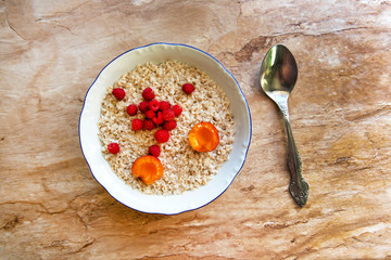 Oatmeal porridge with raspberries and apricots in bowl with blue border on stone table. Top view. Delicious, healthy breakfast.