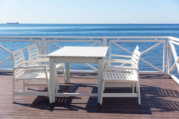 Four white, wooden chairs and a table stand on the wooden flooring in the cafe on the veranda near the blue sea in Sunny weather in summer.
