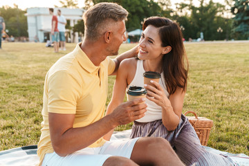 Portrait of happy middle-aged couple drinking coffee takeaway from paper cup while sitting on grass in park
