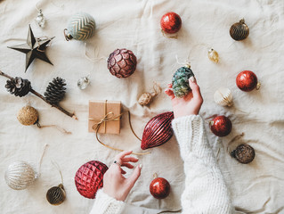 Christmas or New Year background, texture, wallpaper. Flat-lay of Christmas festive decoration toys and woman's hands with pine cone over beige linen cloth, top view. Preparation for winter holidays