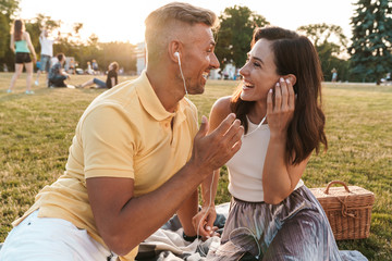 Obraz premium Portrait of excited middle-aged couple man and woman laughing and listening to music together with earphones in summer park