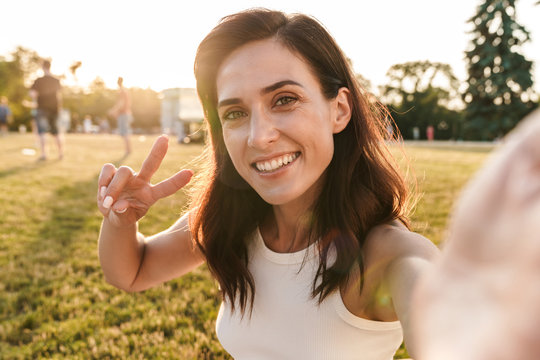 Image of happy middle-aged woman showing peace sign and taking selfie photo in summer park