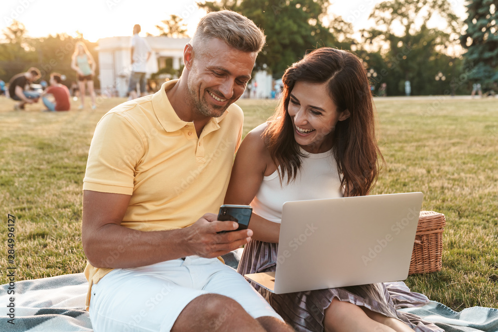 Wall mural portrait of smiling middle-aged couple holding cellphone while using laptop computer during picnic i