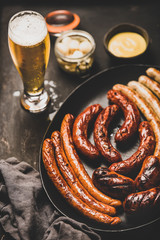 Octoberfest dinner table concept with grilled veal and pork sausages, sauce in jar, pickled vaggies and lager beer in glass over dark background, selective focus, close-up