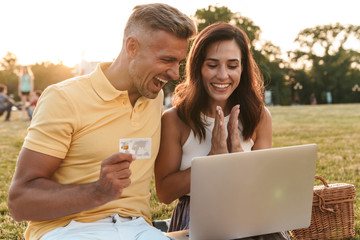 Portrait of happy middle-aged couple holding credit card while using laptop computer during picnic in summer park