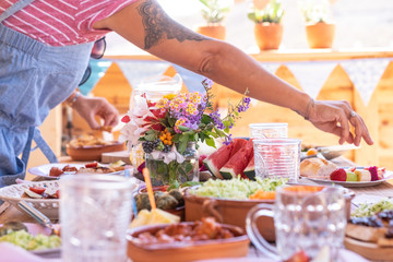 Female arm with black tattoo taking some fruit from the table. Caucasian peoples enjoying brunch or meal together. Fruits and vegetables on the wooden table. Sunlight outdoor on the terrace