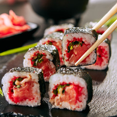 Chopstick holding smoked salmon sushi rolls with green onions, flying fish red caviar, spicy sauce and nori on blurred background. Fresh gourmet japanese food on black slate plate background closeup