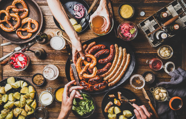 Flat-lay of Octoberfest party dinner table with grilled meat sausages, pretzel pastry, potatoes, cucumber salad, sauces, beers and peoples hands holding food over dark wooden background, top view