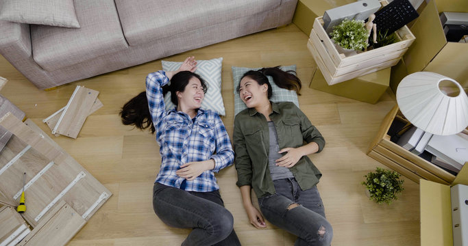 top view two young happy lesbian couple moving into new house. girls lovers lying down on pillows on floor after brought boxes with things relocation apartment. women having fun laugh look each other