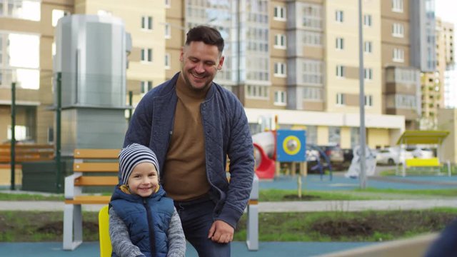 Medium shot of laughing 30-something Caucasian man having fun on playground of urban residential compound on autumn evening with happy 4-year-old son, and helping him play on teeter-totter