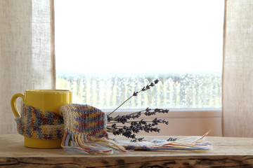 yellow mug in a knitted scarf and a bouquet of lavender on the background of a window after rain. warming fragrant atmosphere of comfort