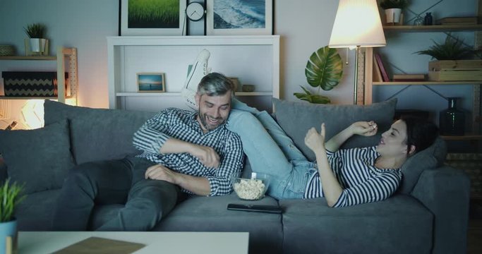 Husband and wife are having fun throwing popcorn laughing watching TV at night on couch in dark apartment. Happy people, joy and entertainment concept.