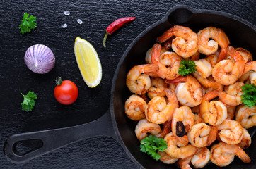tasty roasted shrimps in a cast iron pan with parsley and garlic, chili, tomato with lemon on black background