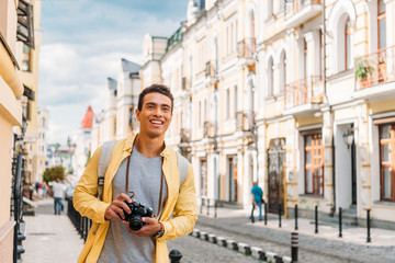 cheerful mixed race man smiling while holding digital camera near buildings