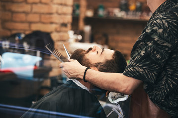 Bearded male sitting in an armchair in a barber shop while hairdresser trip his beard with...