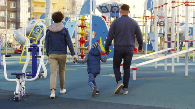 Rear follow shot of mother, father and young boy holding hands together and walking towards outdoor playground at residential compound, parents chatting and child skipping with excitement