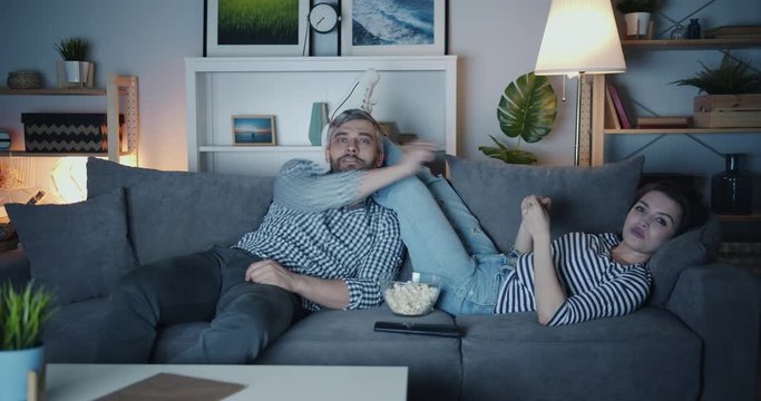Joyful man and woman are watcing TV throwing popcorn and laughing having fun together in house at night. Happy people, relationship and home concept.