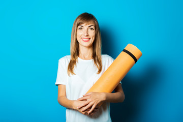 A young woman in a white T-shirt holds a yoga mat on a blue background. The concept of yoga, yoga for beginners, fitness, stretching