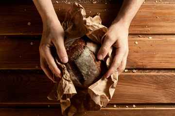 Male hands hold fresh rye bread in a paper bag on the kitchen table with breadcrumbs and grains. Wooden background.